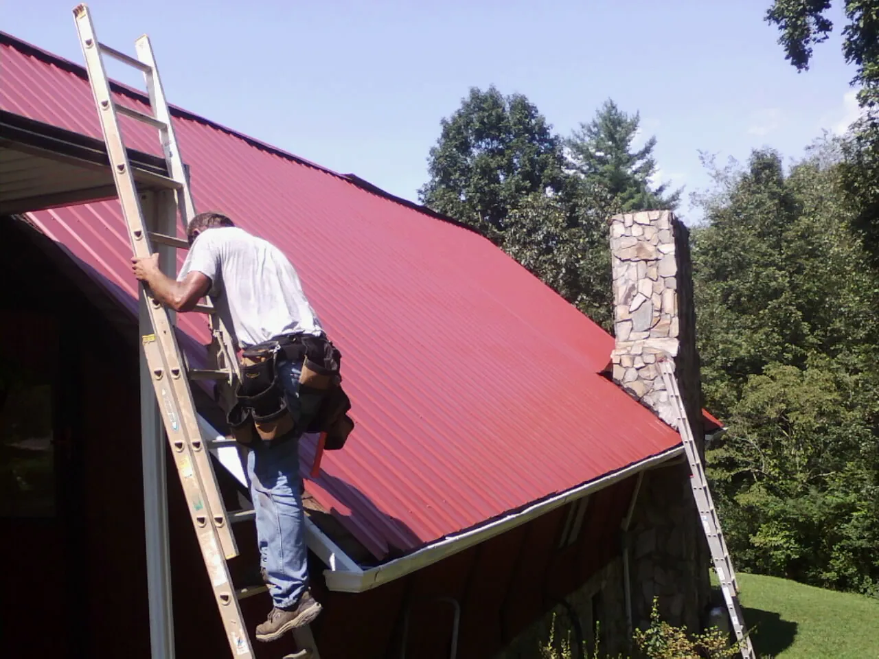 Roofer on a ladder beside a red metal roof and stone chimney — Soffit & Fascia Repair by Roofing Brothers in Myrtle Point